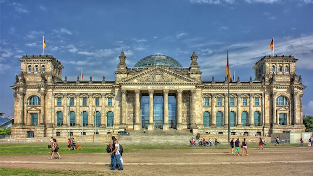 Front view of the Reichstag Building in Berlin with people and flags, under a bright sky.