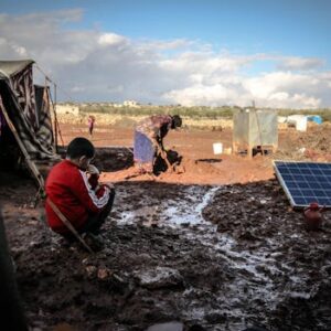 A boy and woman in a muddy refugee camp in Idlib, Syria with tents and solar panel.