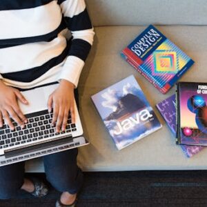 Woman using laptop on sofa, surrounded by programming books, learning coding.