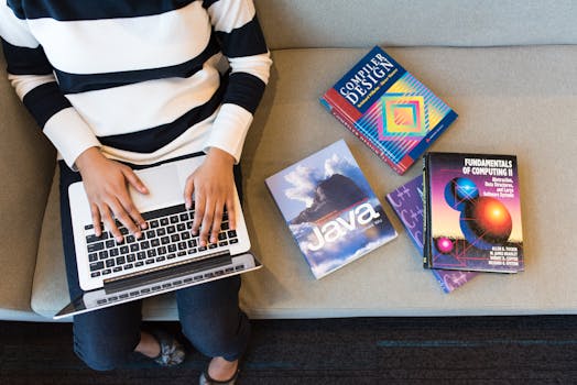 Woman using laptop on sofa, surrounded by programming books, learning coding.