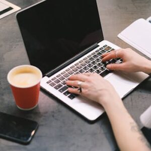 A freelancer typing on a laptop with a cup of coffee, smartphone, and notepad on the table.