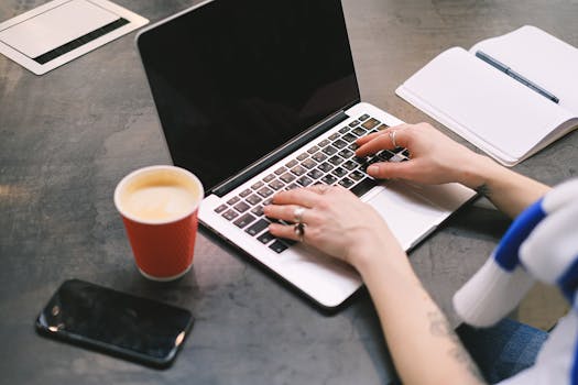 A freelancer typing on a laptop with a cup of coffee, smartphone, and notepad on the table.
