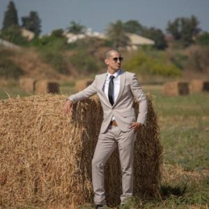 Professional businessman in a suit leans casually on a hay bale in a countryside setting.