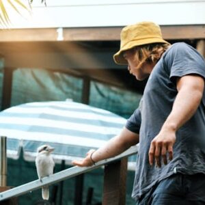 A man wearing a hat interacts with a perched kookaburra outdoors.