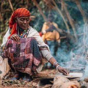 A woman cooking outdoors in a traditional African village setting with visible smoke.