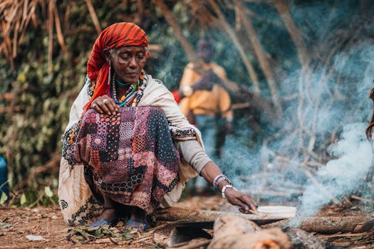 A woman cooking outdoors in a traditional African village setting with visible smoke.