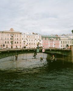 Workers on scaffolding under a historical bridge over a river in an urban setting with classic buildings in the background.