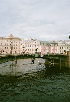 Workers on scaffolding under a historical bridge over a river in an urban setting with classic buildings in the background.