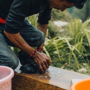 A man kneels by a stream, handwashing clothes with natural surroundings.