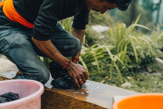 A man kneels by a stream, handwashing clothes with natural surroundings.