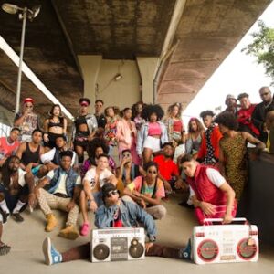 Energetic group posing stylishly under an urban bridge with boomboxes, featuring diverse fashion and expressions.