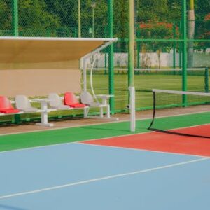 Vibrant tennis court featuring colorful flooring, seating benches, and lush greenery, captured outdoors in daylight.