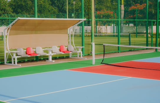 Vibrant tennis court featuring colorful flooring, seating benches, and lush greenery, captured outdoors in daylight.