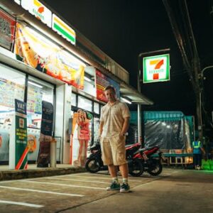 A man stands outside a brightly lit 7-Eleven store at night in Phuket, Thailand.