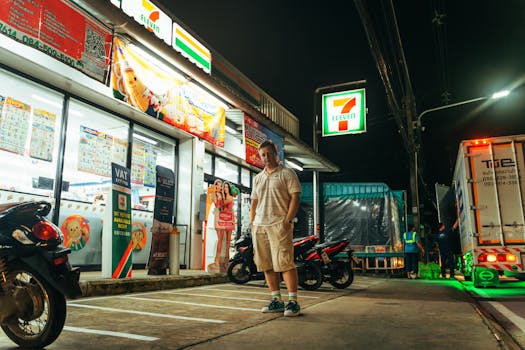 A man stands outside a brightly lit 7-Eleven store at night in Phuket, Thailand.