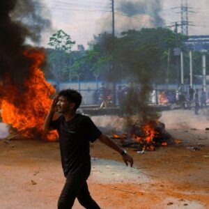 A man walks past burning debris during an outdoor protest in a city, reflecting tension and unrest.