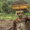A determined rural man carries a basin of soil on his head through lush countryside.