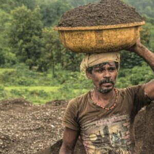 A determined rural man carries a basin of soil on his head through lush countryside.