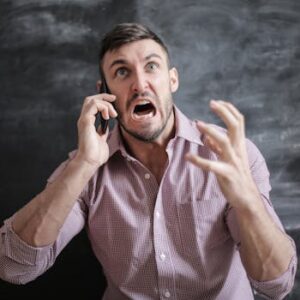 A distressed man in a pink shirt expressing frustration while using a smartphone indoors.