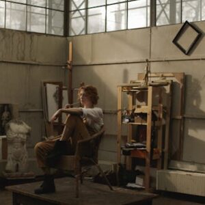 Artist in a chair reflecting in an industrial-style studio filled with sketches and art materials.