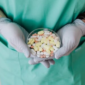 Crop male doctor wearing medical uniform and latex gloves holding glass bowl with colorful different drugs with crossed hands during work