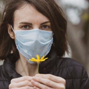 A woman wearing a surgical mask holds a single yellow flower outdoors, promoting health and safety.