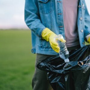 Volunteer in denim jacket and gloves picks up plastic bottle into trash bag in green field.