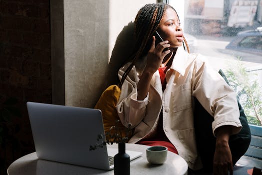 Woman with braids talking on phone in a cafe, sunlight through window.