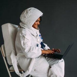 Smiling astronaut in white space suit sits indoors using a laptop, showcasing technology and exploration.