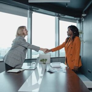 Two businesswomen shaking hands in a modern office setting, symbolizing partnership and agreement.