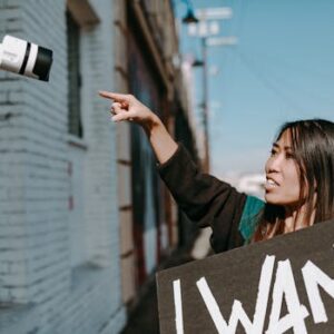 A woman holds a placard, pointing at a CCTV camera during an outdoor protest.