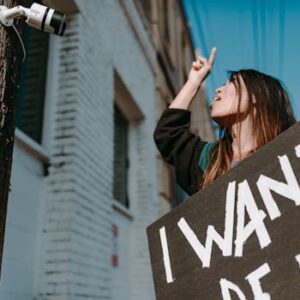A woman gestures toward a CCTV camera while holding a protest sign in a city setting.