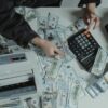 Hands counting 100 dollar bills using a calculator and money counter on a table.
