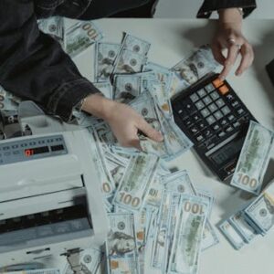 Hands counting 100 dollar bills using a calculator and money counter on a table.