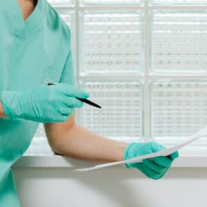 A healthcare worker in scrubs and gloves holding a pen and paper in a medical facility.