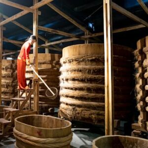 Woman in red clothing overseeing fermentation process in a traditional wooden barrel warehouse.