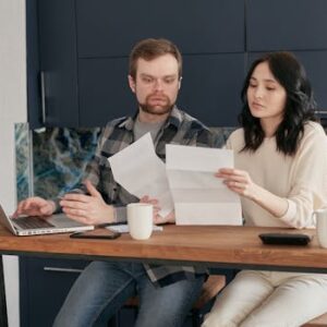 A couple sitting at a wooden table, reviewing documents with a laptop nearby, expressing concern.