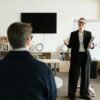 Businesswoman giving presentation in office with bookshelves and clock.