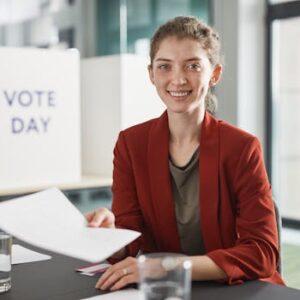 Cheerful young woman sitting indoors, holding documents during vote day with a smile.