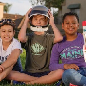 Three kids sitting on grass, playing with a toy rocket, enjoying a sunny day.