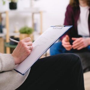 Close-up of a therapist writing notes on a clipboard while conversing with a patient.