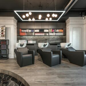 Black leather backwash chairs with sinks under glowing lamps placed in modern spacious beauty salon with cosmetology products and window