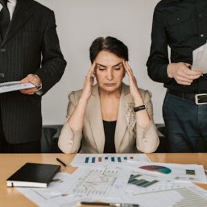 A stressed businesswoman holding her head with colleagues holding documents in a corporate office setting.