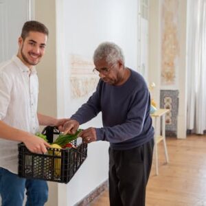 A young volunteer offers support by delivering fresh vegetables to an elderly gentleman indoors.
