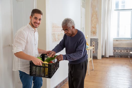 A young volunteer offers support by delivering fresh vegetables to an elderly gentleman indoors.
