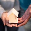 Hands holding a small wooden house model, symbolizing new home ownership and real estate transactions.