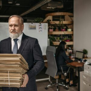 Elderly man in suit holding large pile of documents in a modern office setting.