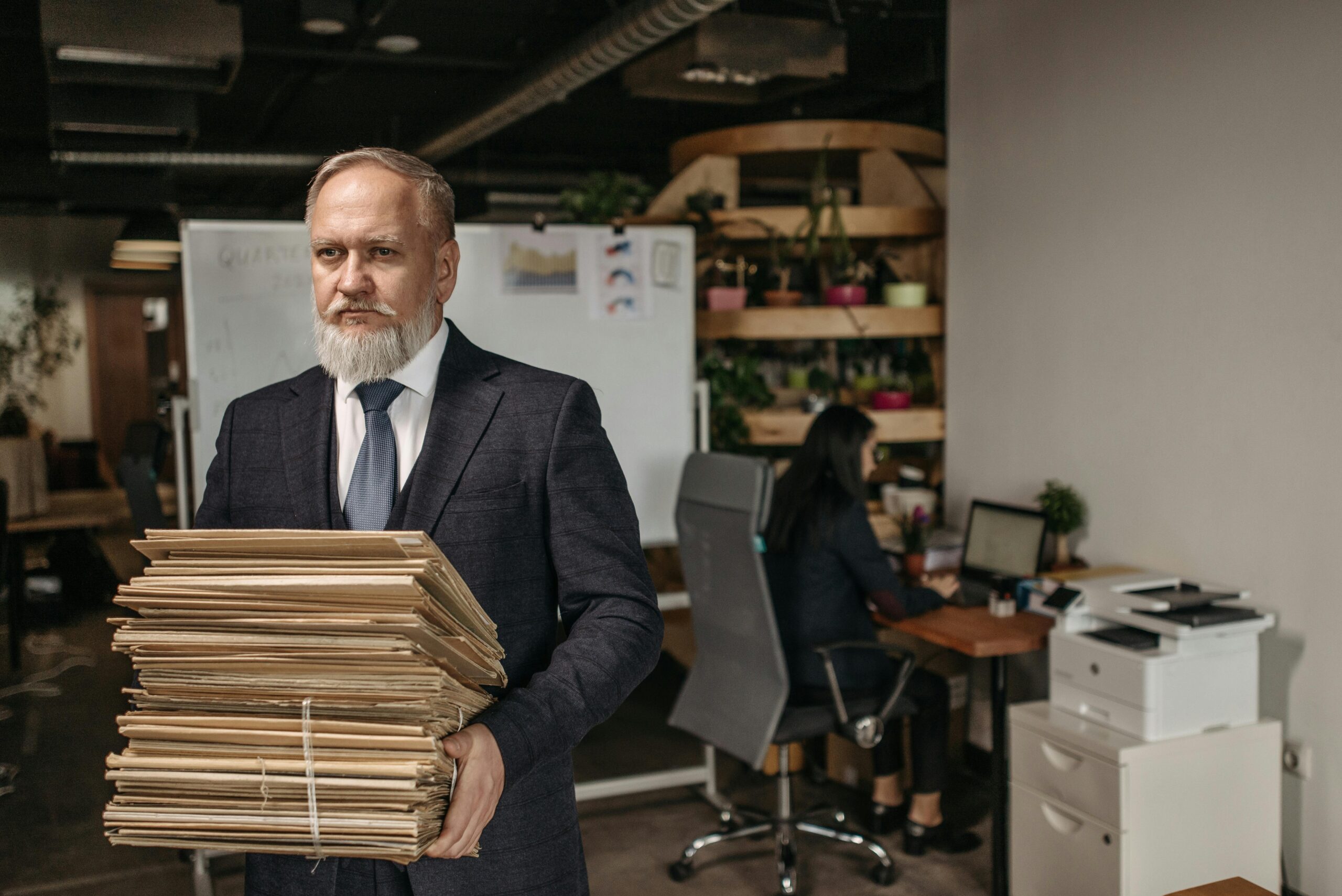 Elderly man in suit holding large pile of documents in a modern office setting.