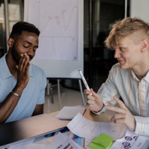 Two young men collaborating with charts, paper, and a laptop on a desk indoors.