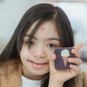 Young girl with Down syndrome smiling and holding a memory game card indoors.